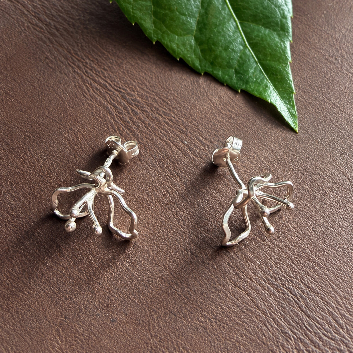 Oktoberblommans handsmidda stiftörhängen i silver på brun bakgrund med grönt blad